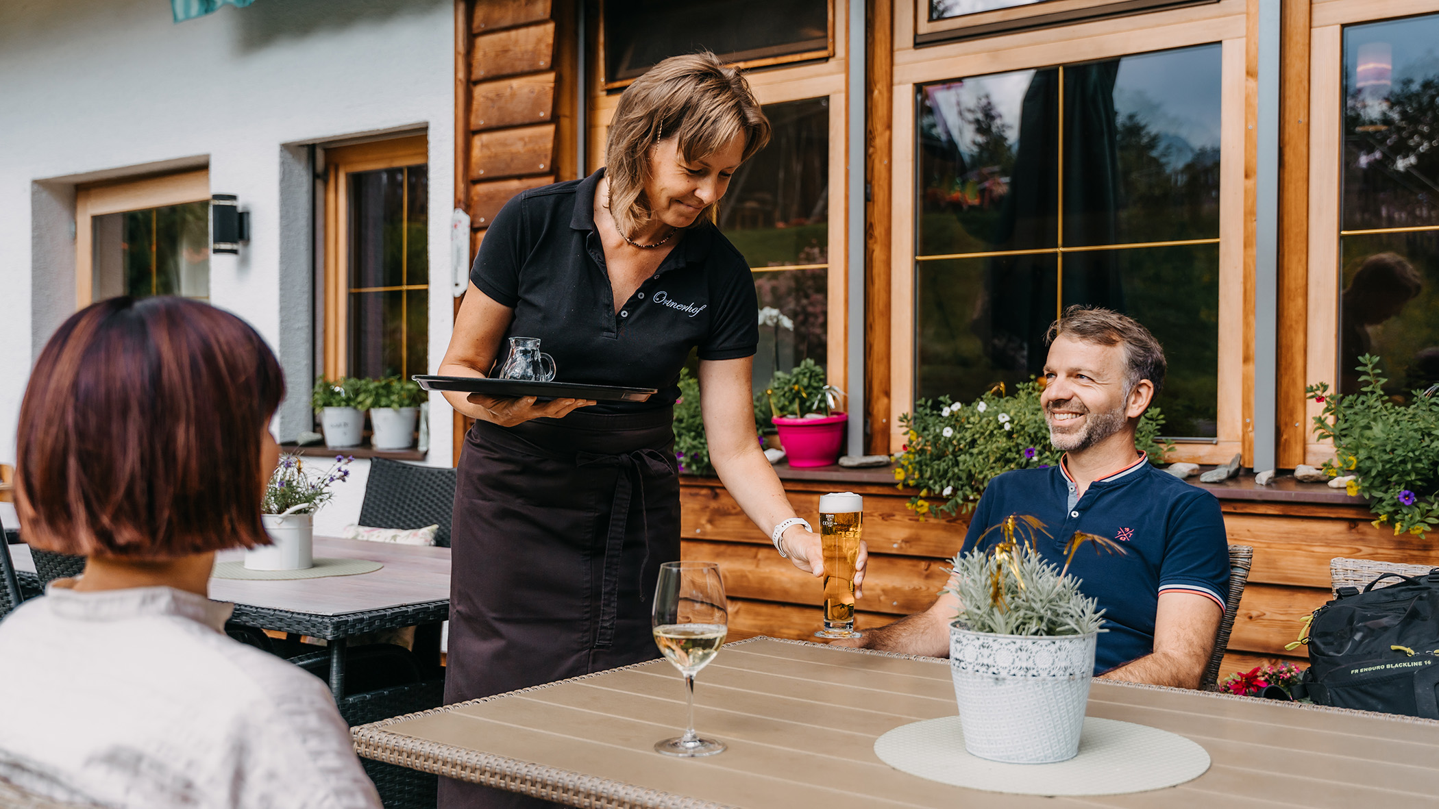 Kellnerin serviert ein Glas Bier und ein Glas Wein an Gäste auf der Terrasse - Hotel Ortnerhof