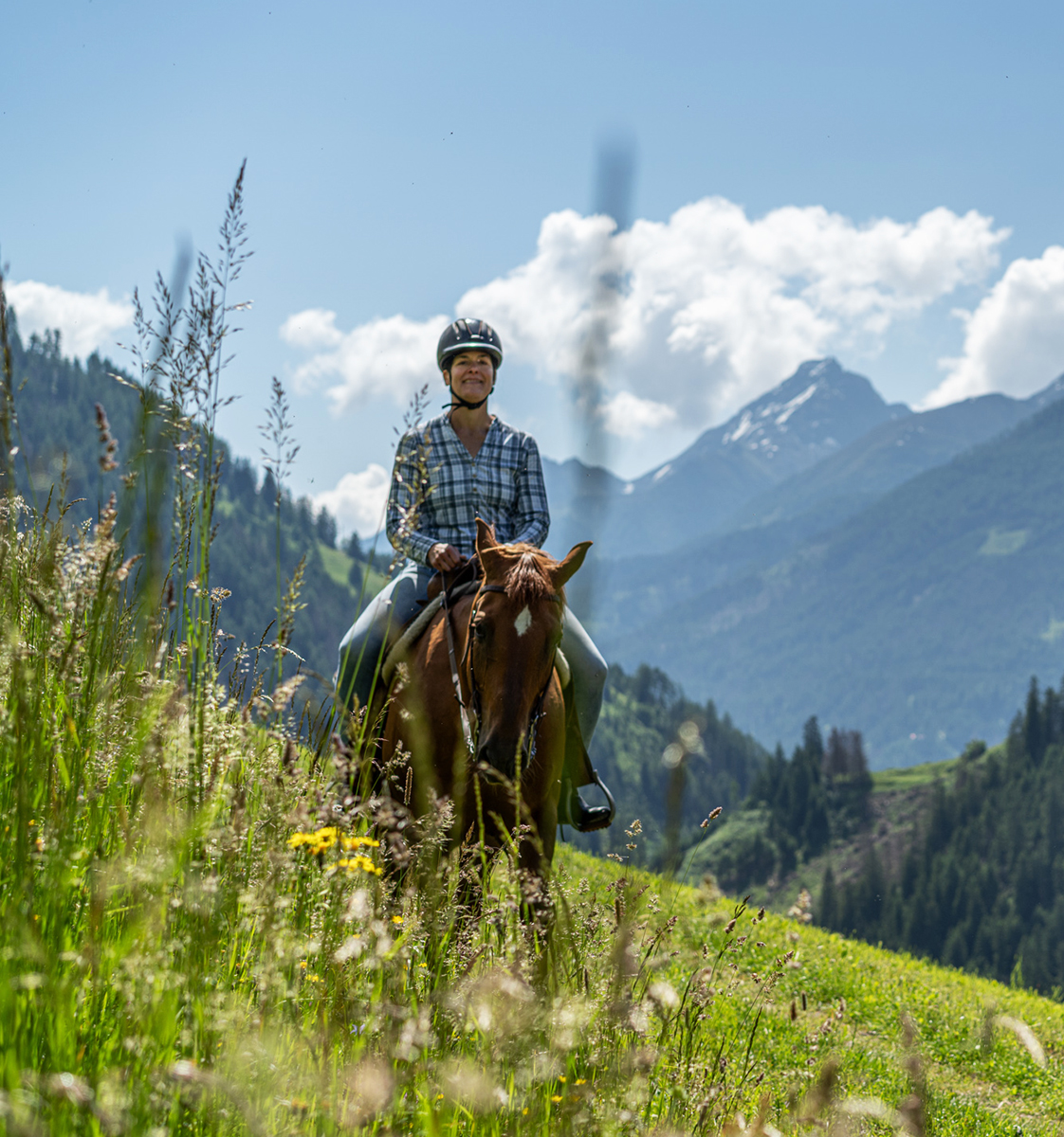 Eine Frau reitet mit lockeren Zügeln durch blühenden Bergwiesen mit grandiosem Bergpanorama im Hintergrunde - Hotel Ortnerhof